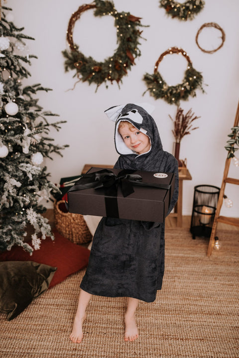 1. Child wearing RÄTT Long-Sleeve Poncho WOLF in dark color, holding a gift box, in a cozy holiday setting