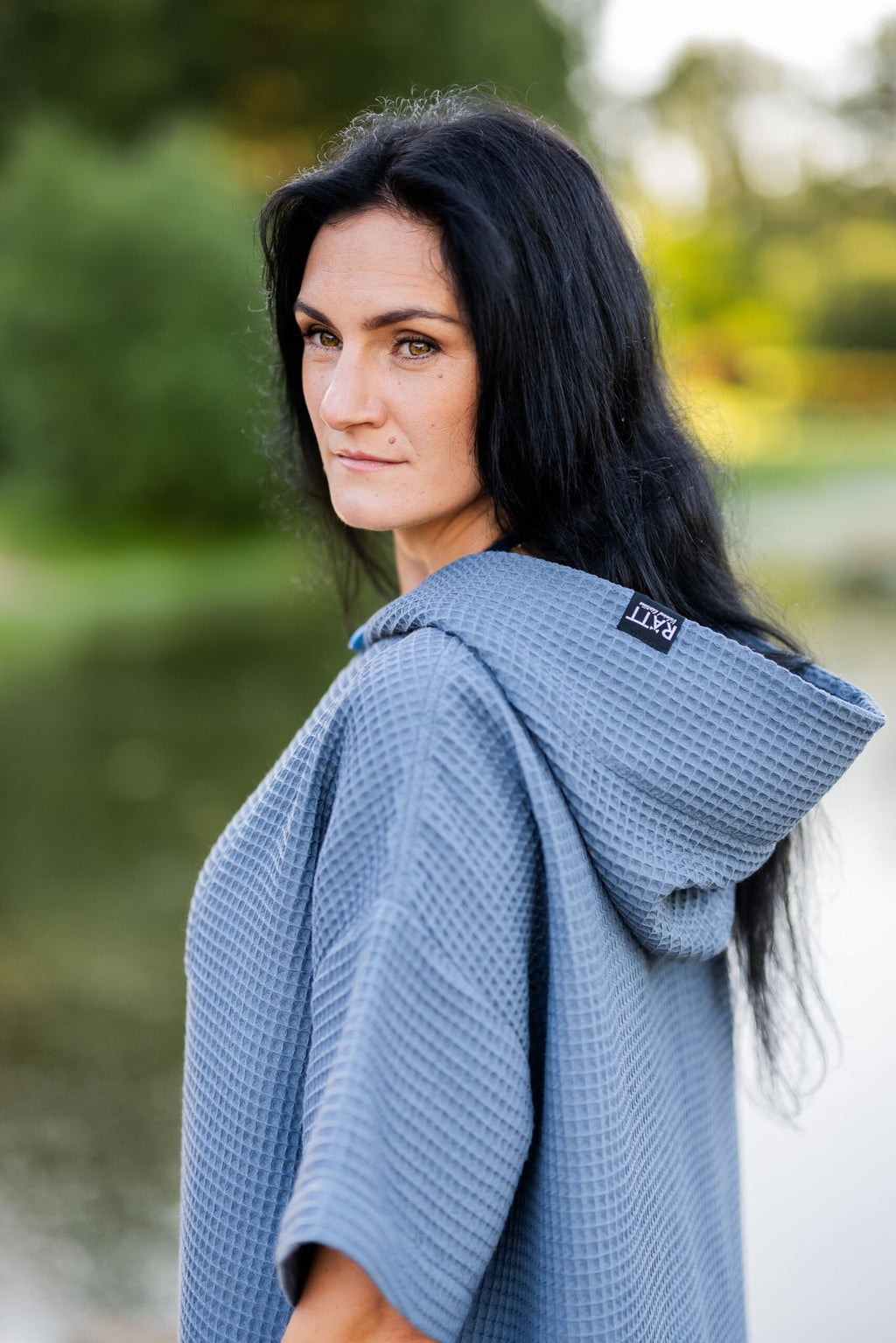 5. Woman in blue RÄTT short-sleeve surf poncho with hood, standing by a lake