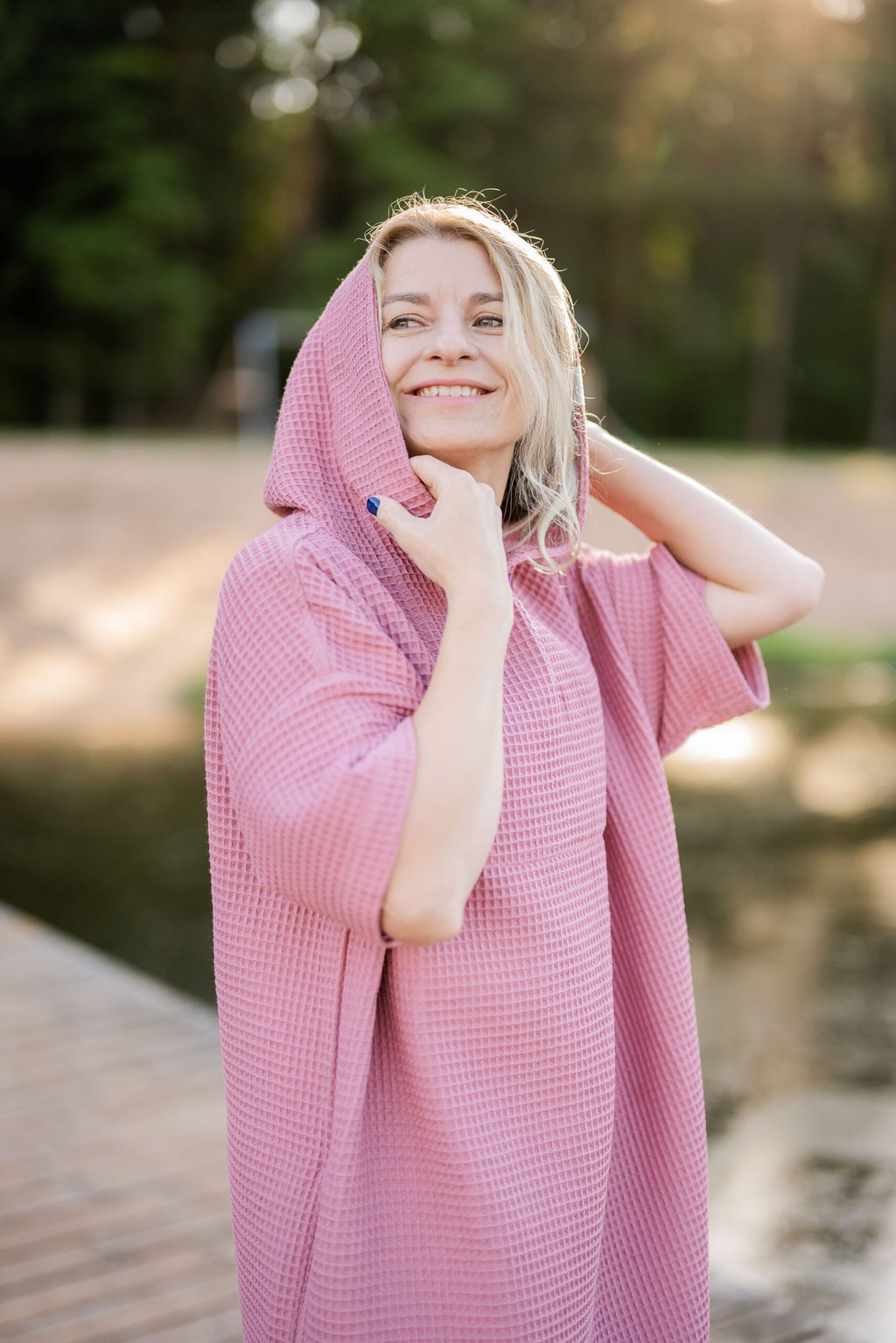 4. Woman wearing pink RÄTT short-sleeve surf poncho with hood up, smiling outdoors