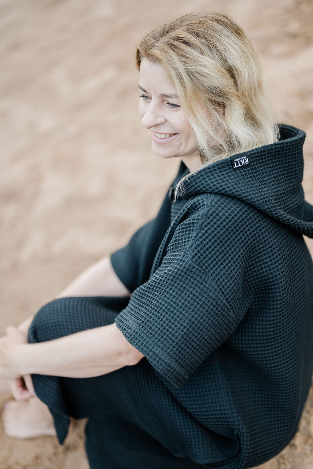 8. Woman sitting on sand wearing black RÄTT short-sleeve surf poncho with hood