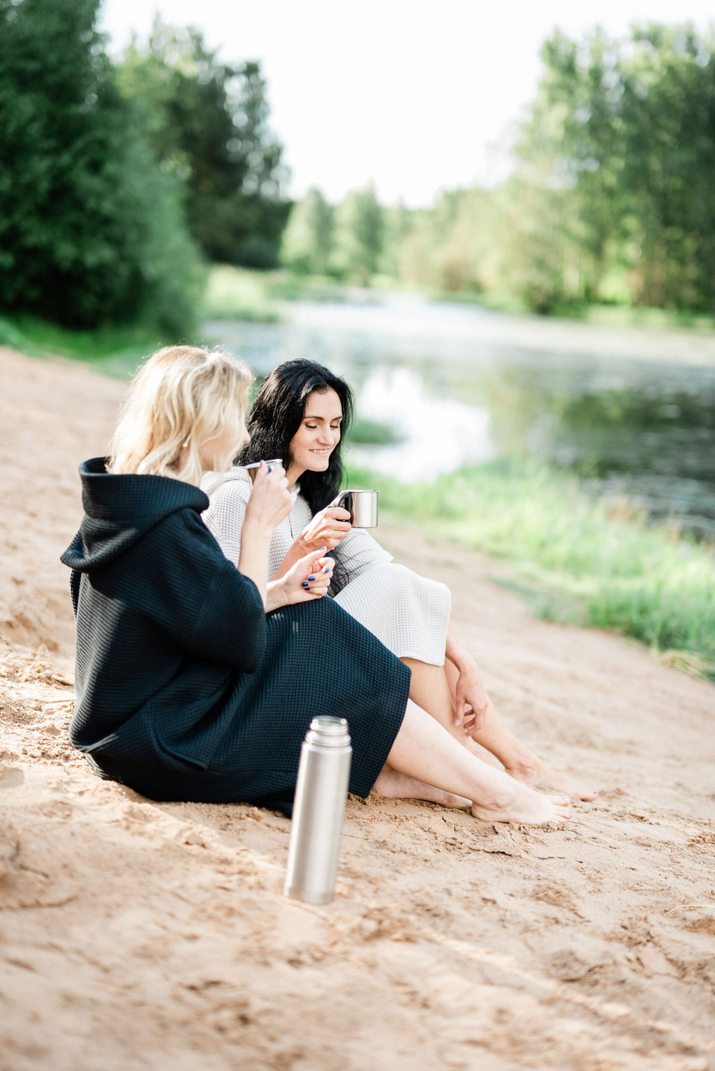 2. Two women sitting on beach wearing RÄTT short-sleeve surf ponchos, enjoying a drink
