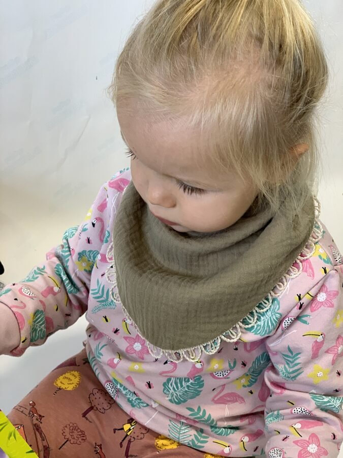3. Child wearing khaki muslin scarf-bib with lace trim, dressed in a pink floral outfit, sitting indoors