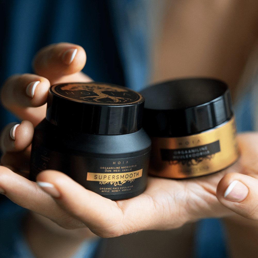 2. Hands holding two jars of HOIA homespa Organic Face Scrub, close-up view