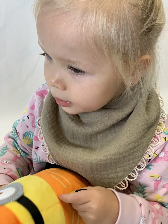 2. Child wearing khaki muslin scarf-bib with lace trim, holding a toy, dressed in a floral outfit
