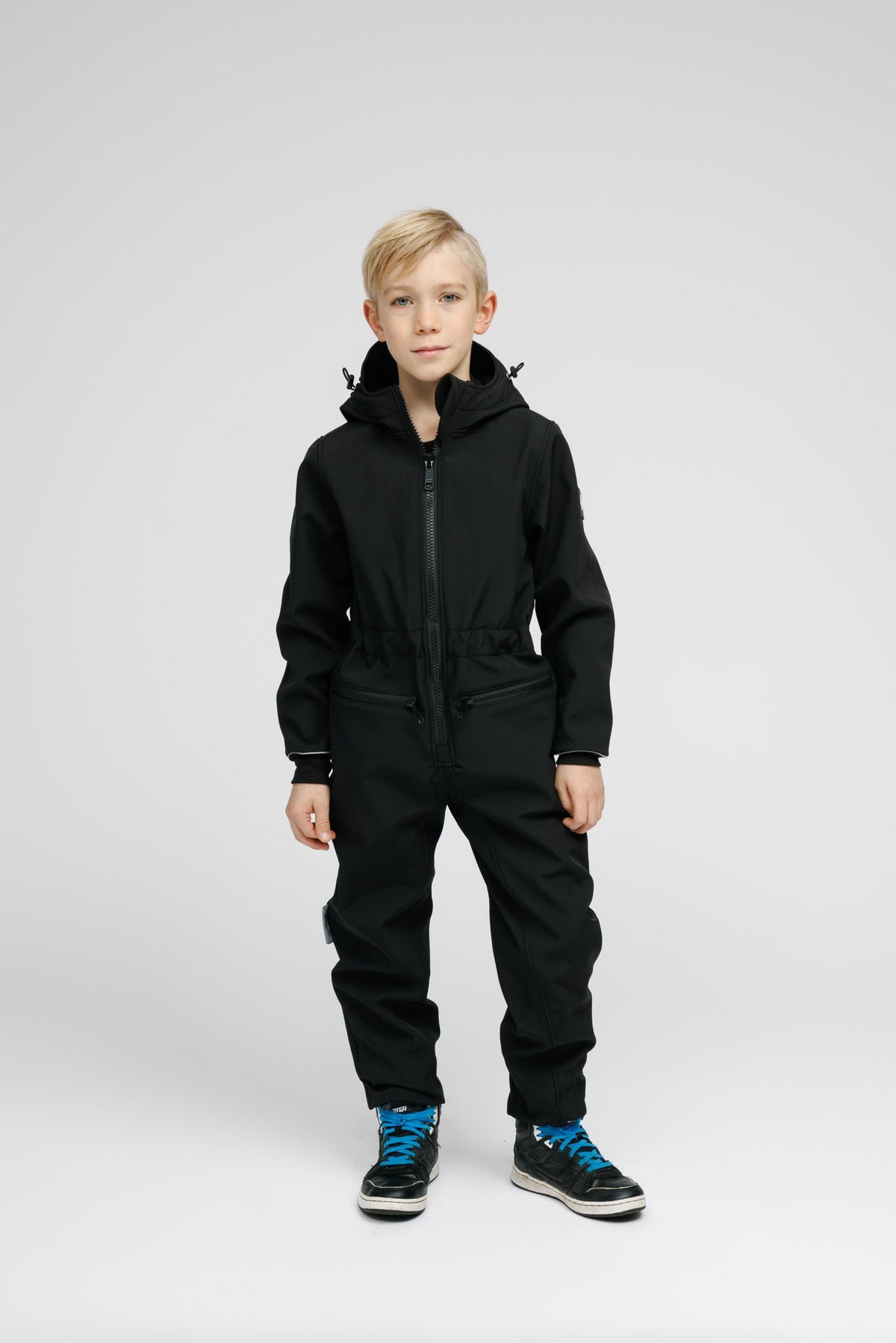 1. Child wearing black softshell onesie with hood and blue sneakers standing in studio setting
