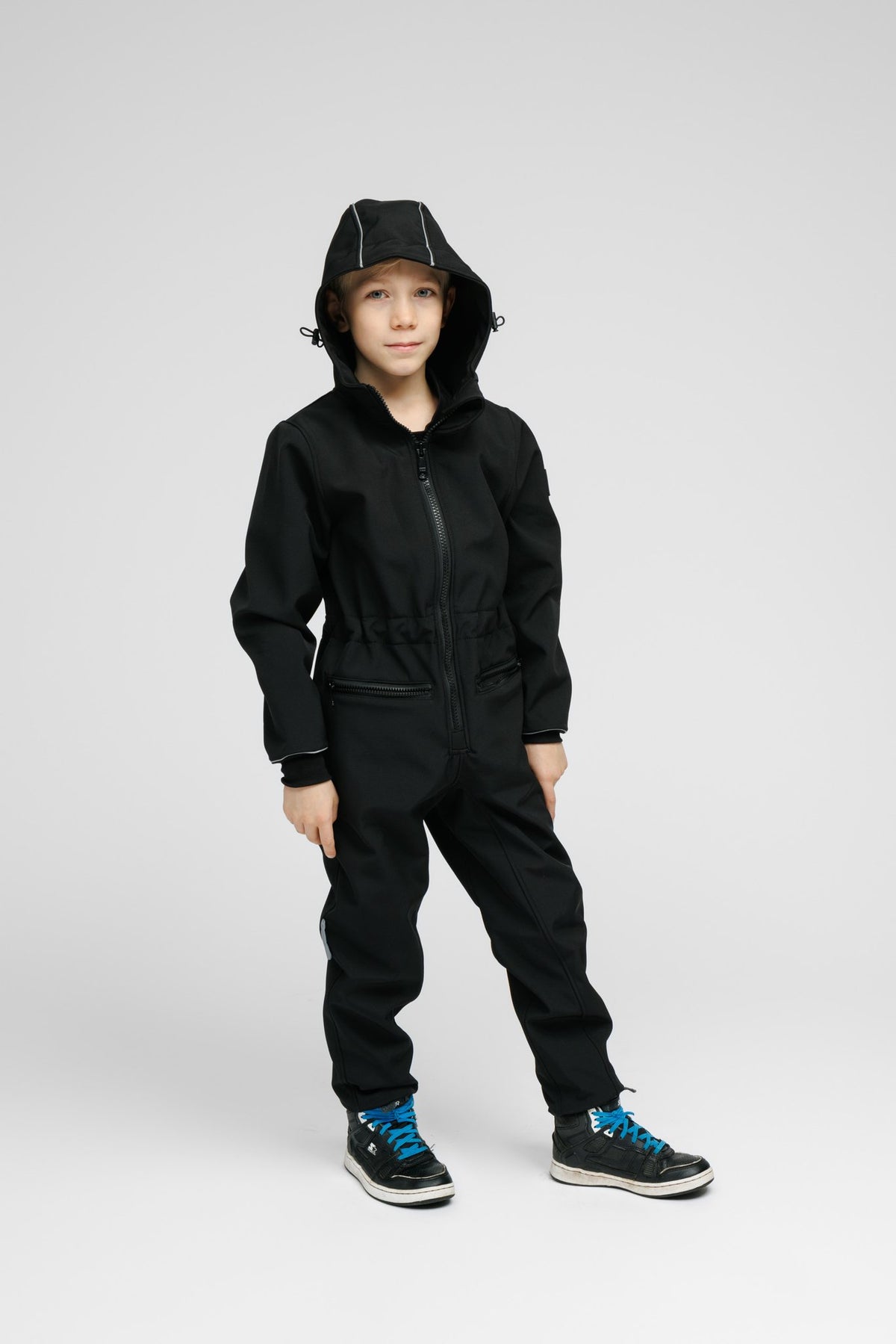4. Child wearing black softshell onesie with hood up and blue sneakers in studio
