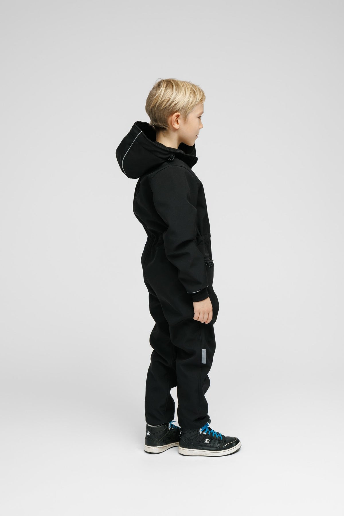 2. Side view of child in black softshell onesie with hood and blue sneakers in studio