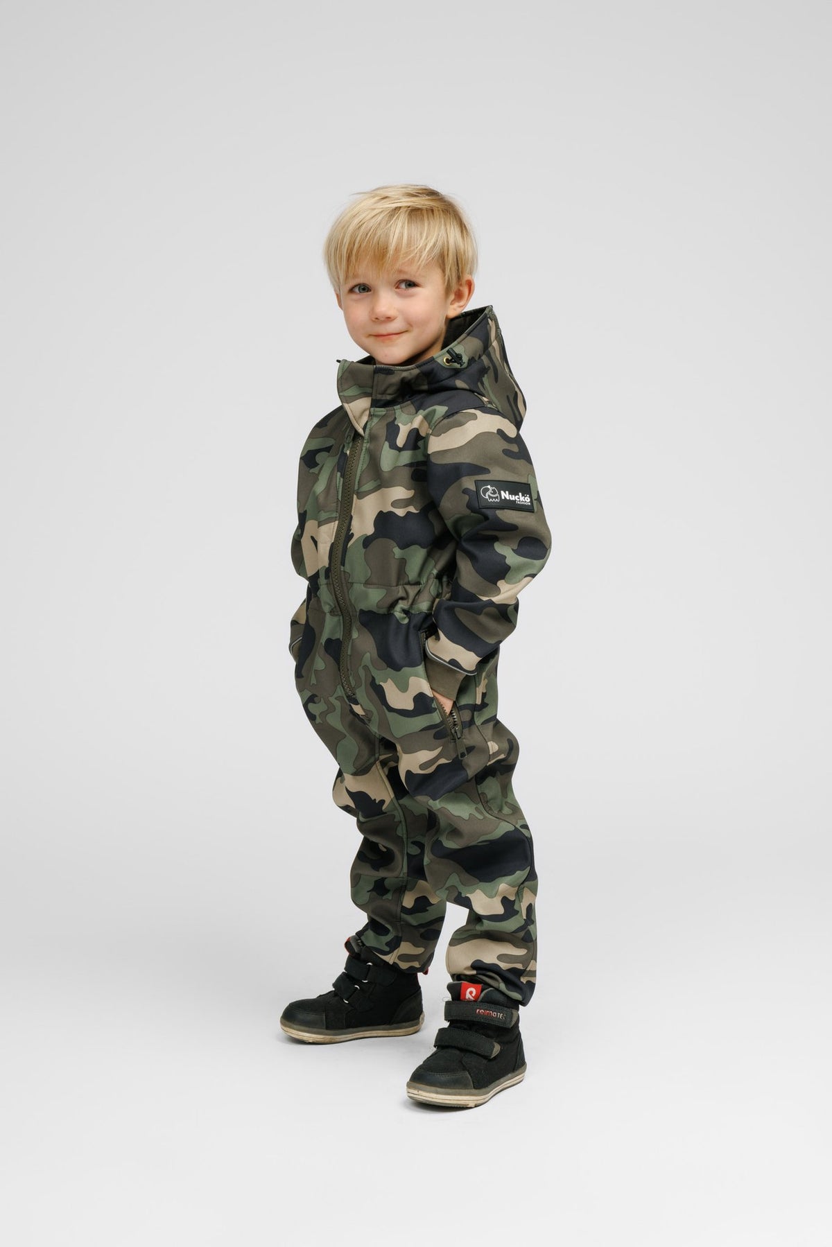 4. Child in Nuckö camouflage softshell onesie, smiling with hands in pockets, displaying brand logo on sleeve, studio background