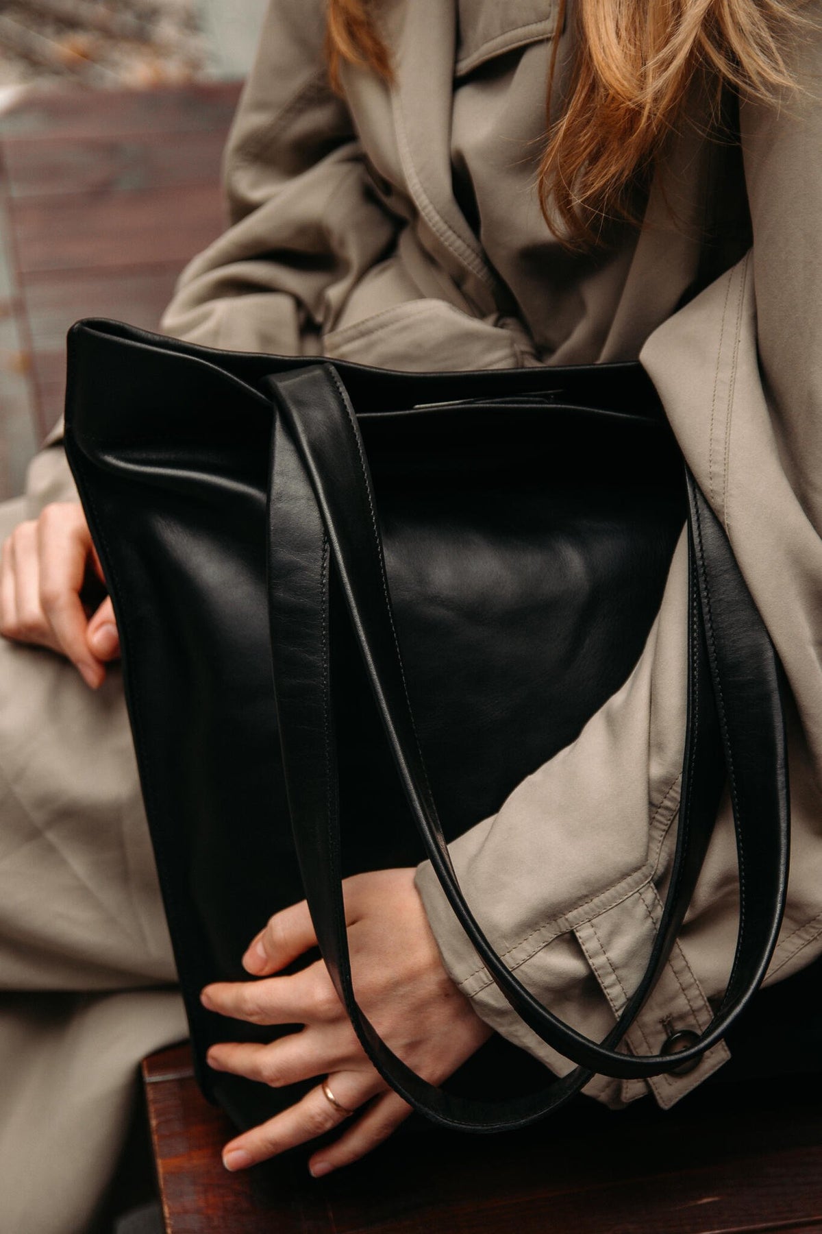 1. Woman holding black leather tote bag by Stella Soomlais, sitting on a bench wearing a beige trench coat