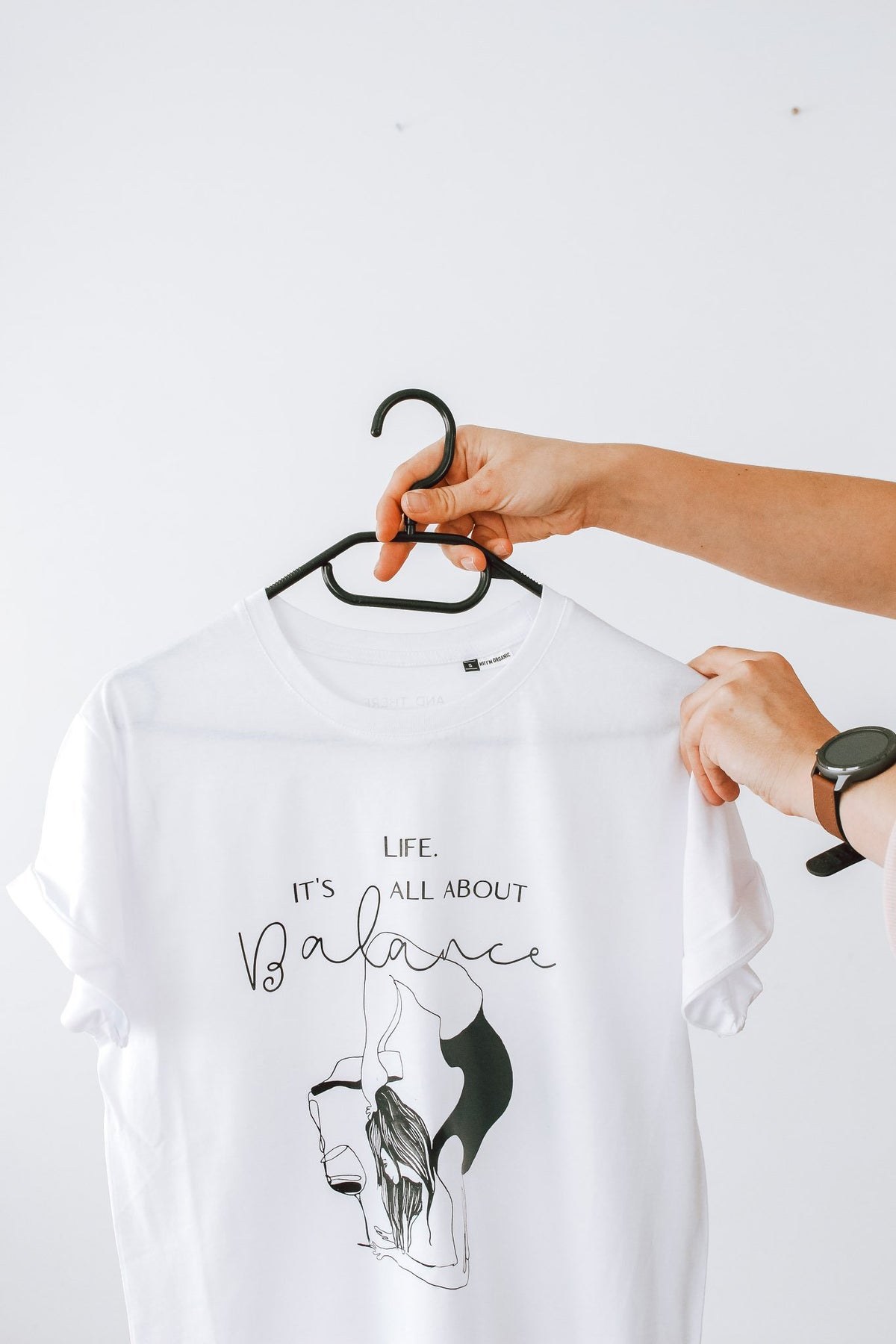 3. Close-up of white 'It's All About Balance' T-shirt by Black Giraffe Brand on hanger, highlighting design and rolled-up sleeves