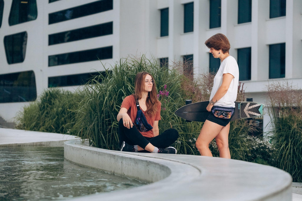 1. Women's lifestyle image featuring Drinbags Classic Waist Bag in COSMIC DOTS pattern, worn by two women outdoors, one holding a skateboard