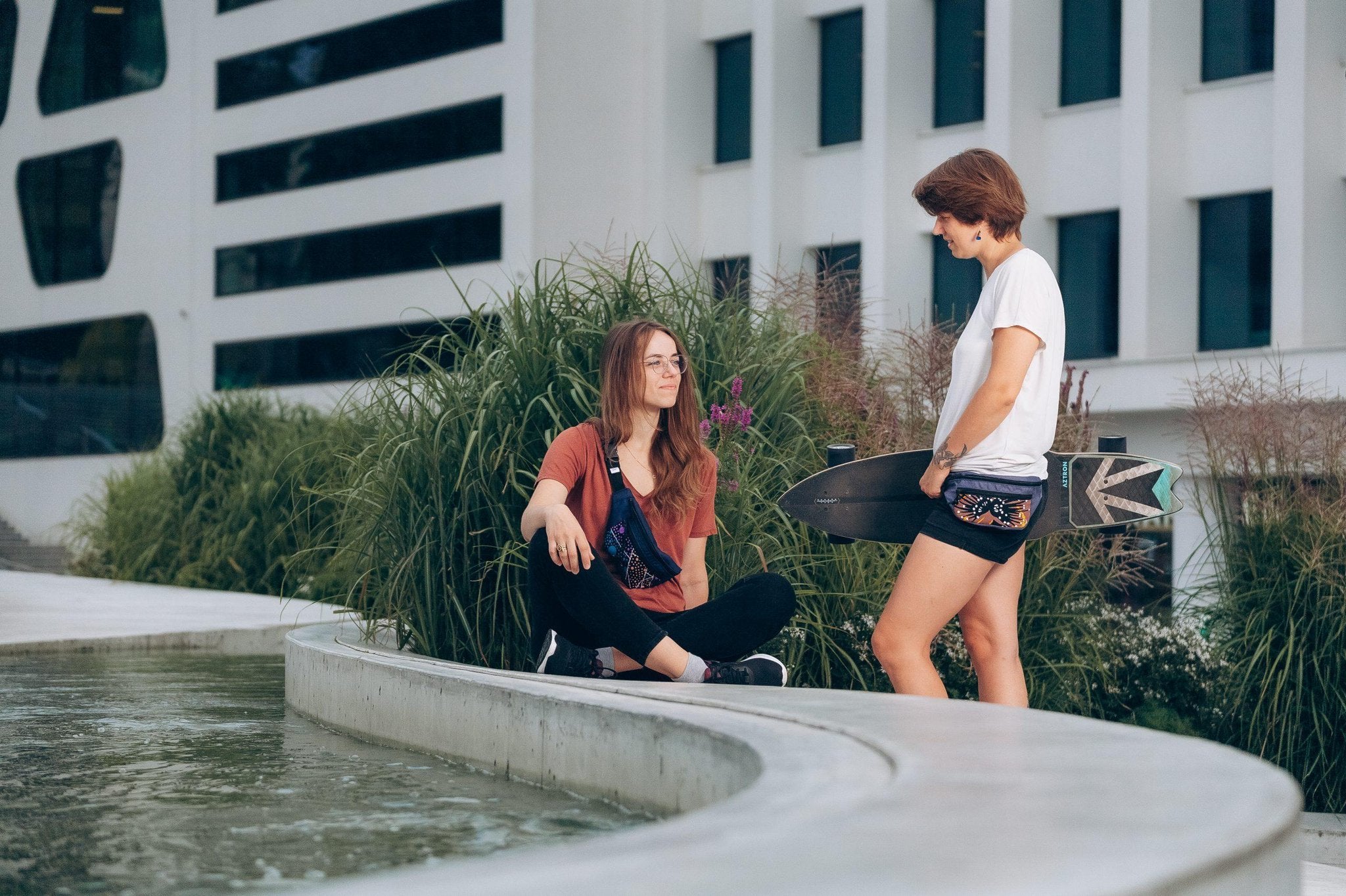 1. Women's lifestyle image featuring Drinbags Classic Waist Bag in COSMIC DOTS pattern, worn by two women outdoors, one holding a skateboard