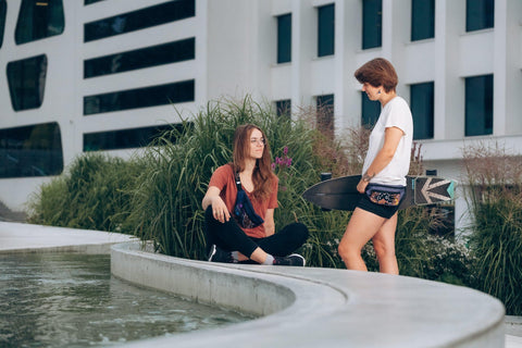 1. Women's lifestyle image featuring Drinbags Classic Waist Bag in COSMIC DOTS pattern, worn by two women outdoors, one holding a skateboard