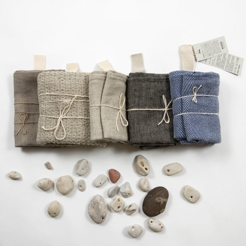 8. Group of folded linen towels in various colors, including blue fishbone, with natural string ties and tags, on white background with stones
