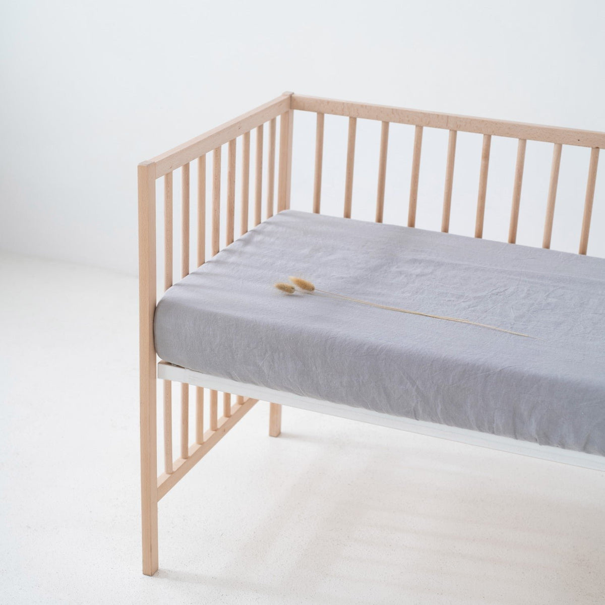 1. Light grey linen fitted sheet on a wooden crib in a minimalist room, showcasing the soft texture and elastic hem