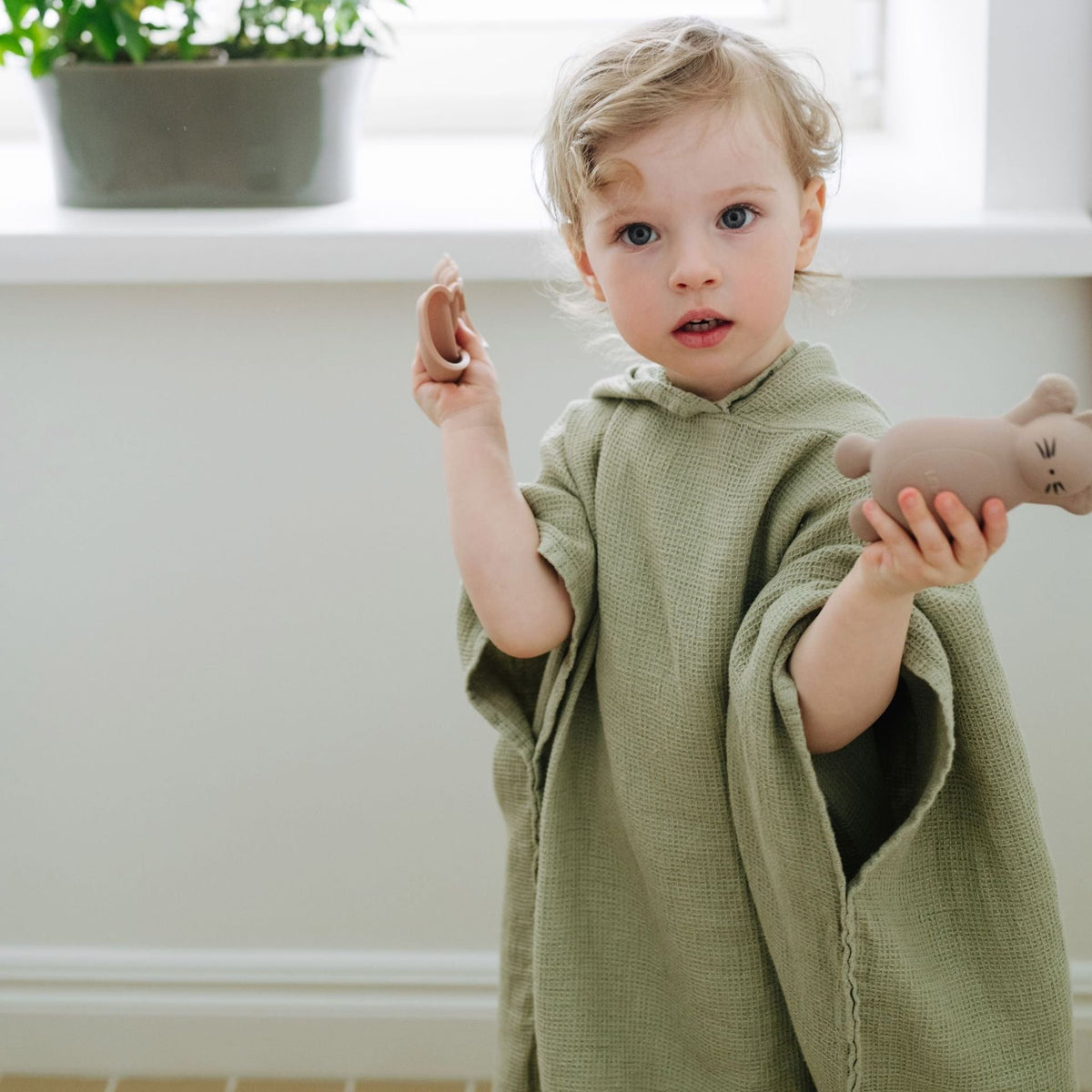1. Child wearing sage green linen poncho with hood, holding toys, standing indoors near a window