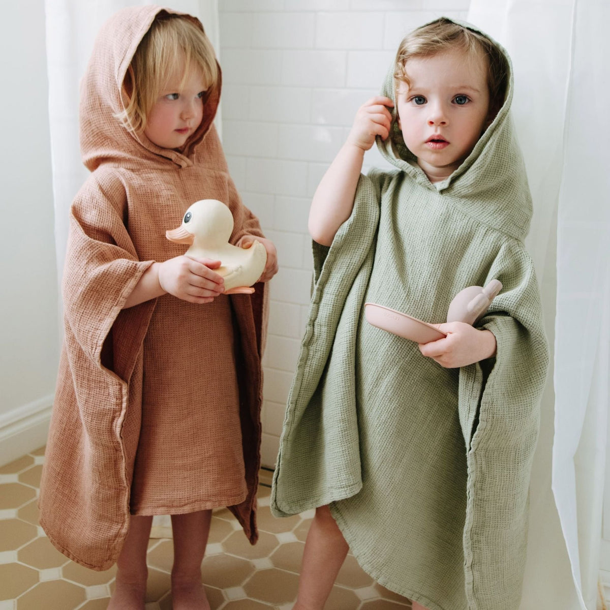 1. Two children wearing linen ponchos, one in sage green and the other in peach, standing in a bathroom holding toys
