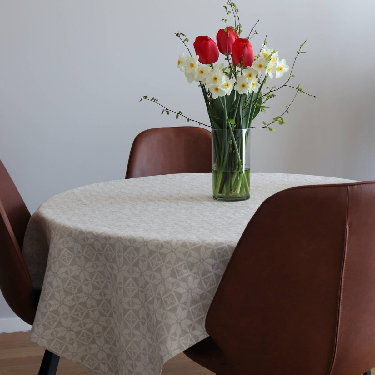1. Beige tablecloth with Hiiumaa kaheksakanna mitten pattern on round table, decorated with vase of red and white flowers, surrounded by brown chairs in a modern dining room setting