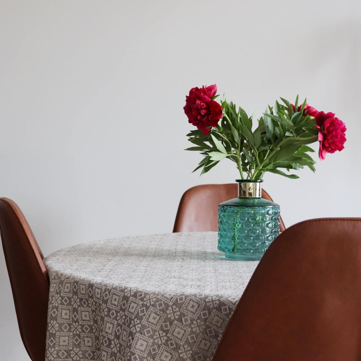 1. Round table with LOKO Tartumaa tablecloth featuring Kodavere folk art pattern, decorated with a blue vase and red flowers, surrounded by brown chairs in a dining room setting