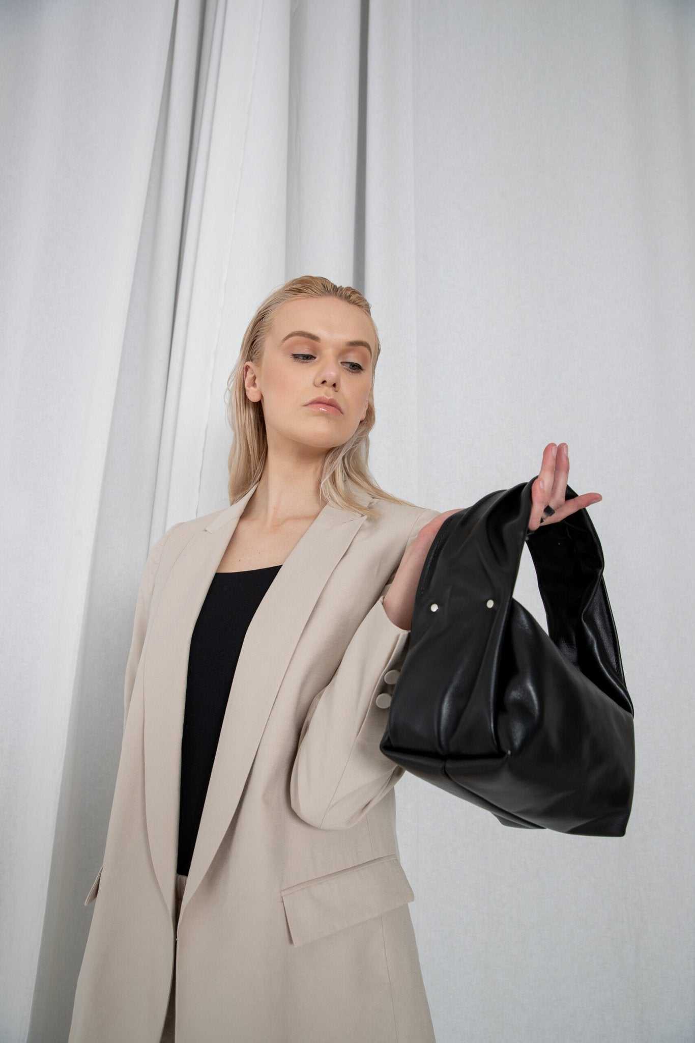 1. Woman holding PYKOK Soft Leather Baguette Handbag Luna in black, styled with beige blazer in studio setting