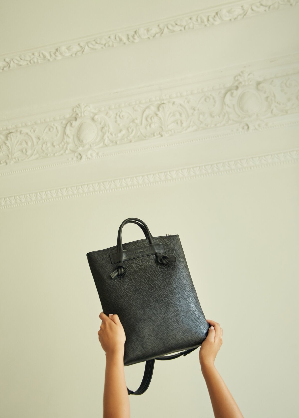 2. Hands holding up Lava Flow ALICE ARM black leather backpack against ornate wall, highlighting knot details