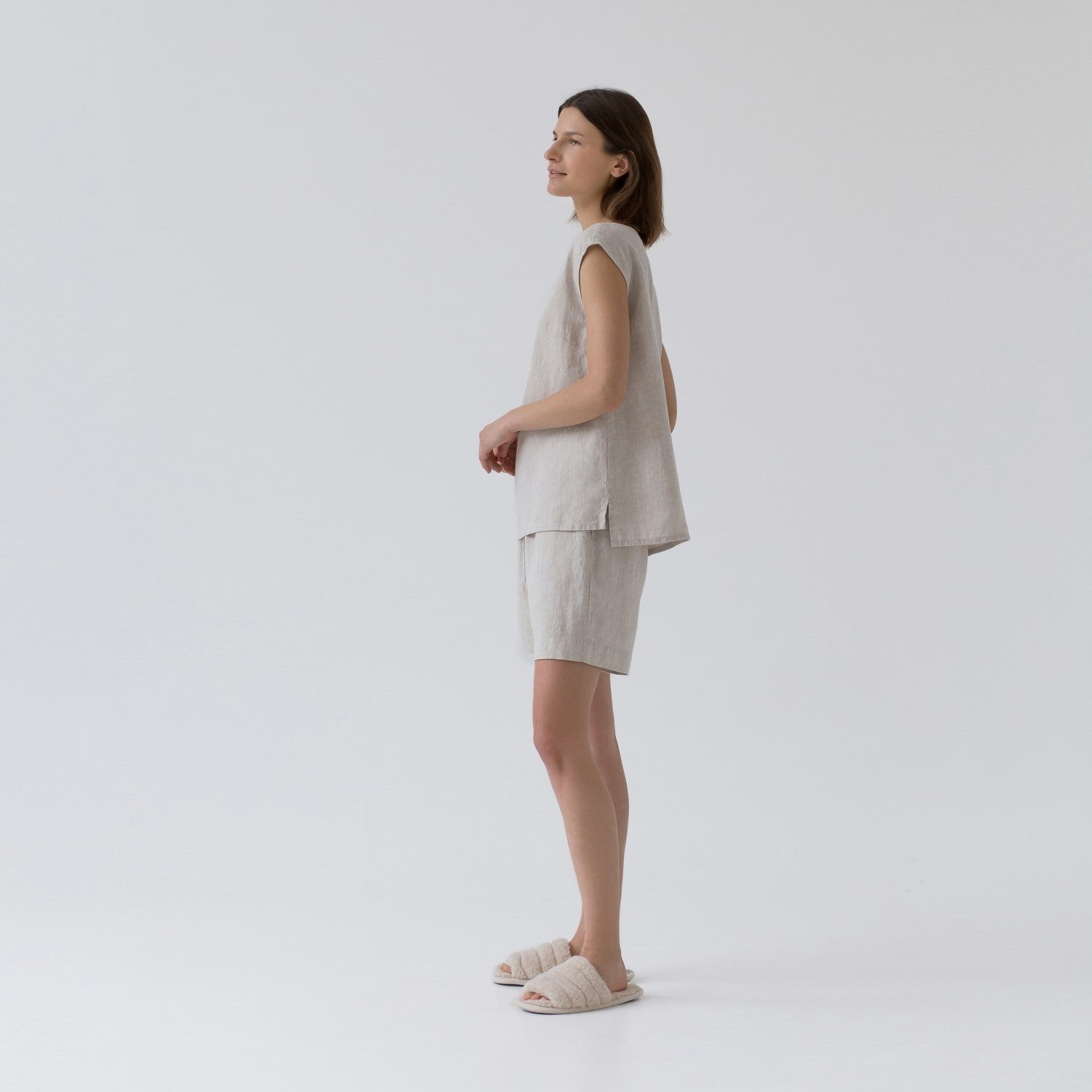1. Woman wearing sleeveless beige linen pyjama set with relaxed fit, standing sideways in a studio setting