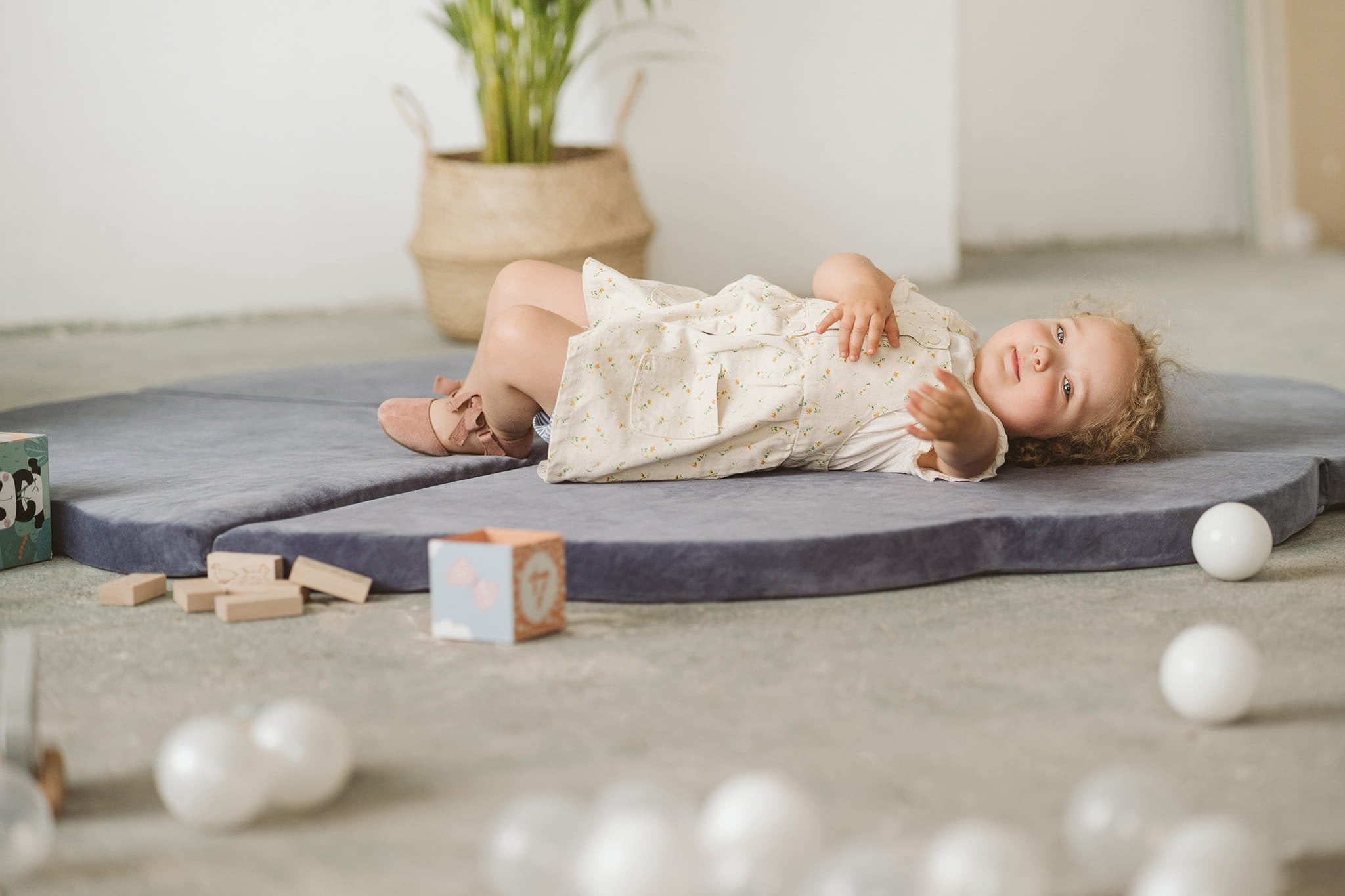 1. Child lying on blue-grey cloud-shaped play mat in a room with toys and plants, showcasing soft velvet texture