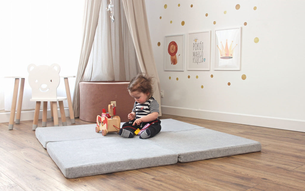 1. Child sitting on square light grey play mat in a decorated room with wooden floor