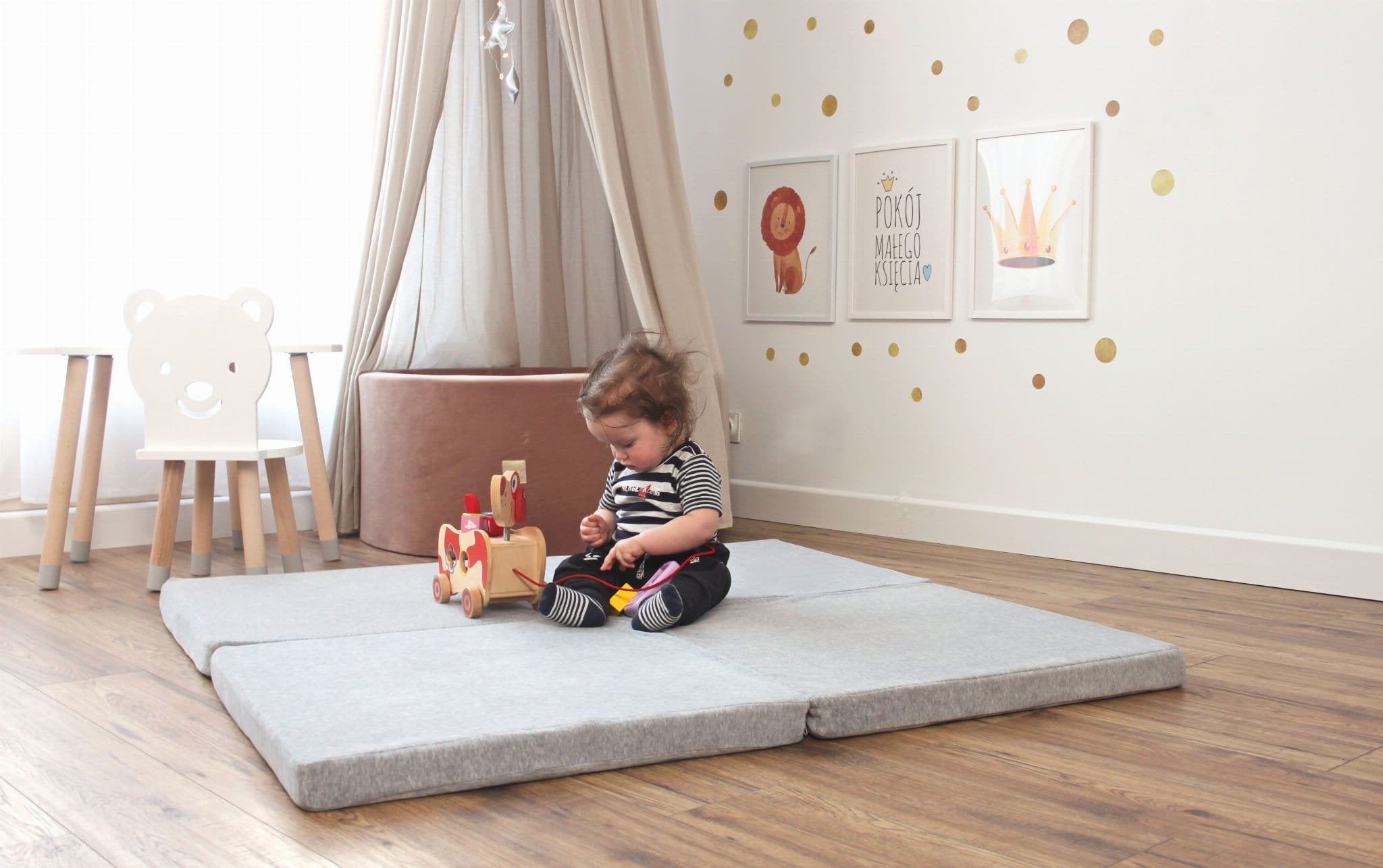 1. Child sitting on square light grey play mat in a decorated room with wooden floor