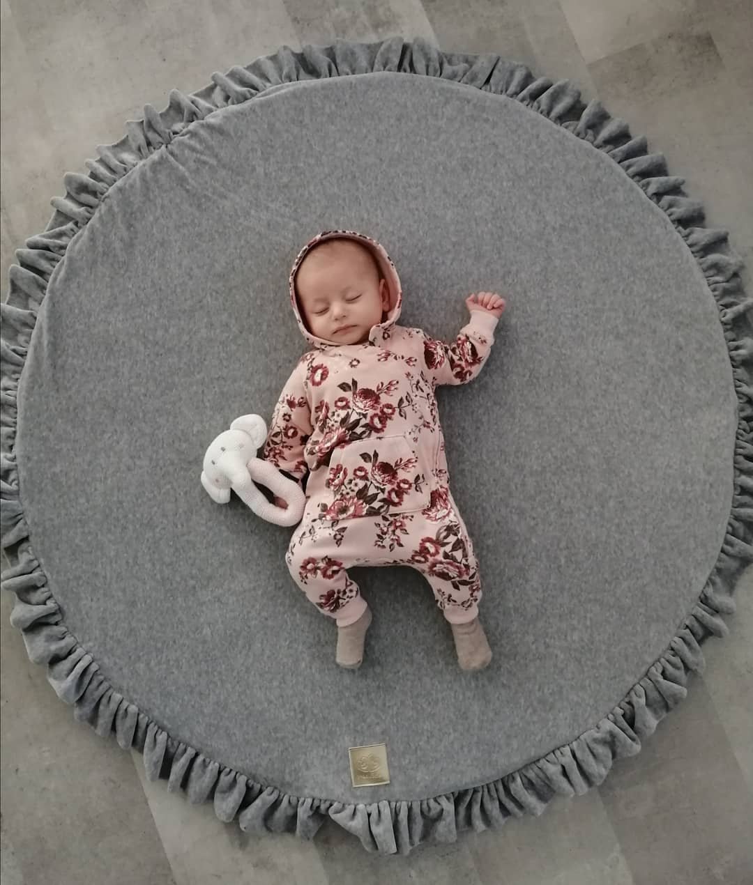 1. Baby sleeping on grey round play mat with frill, wearing floral outfit and holding white toy
