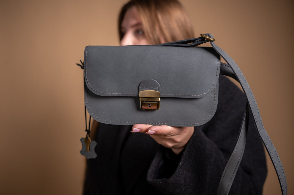 7. Front view of grey leather saddle bag mini by Zelma Kraft, held by woman, highlighting buckle closure and elegant design