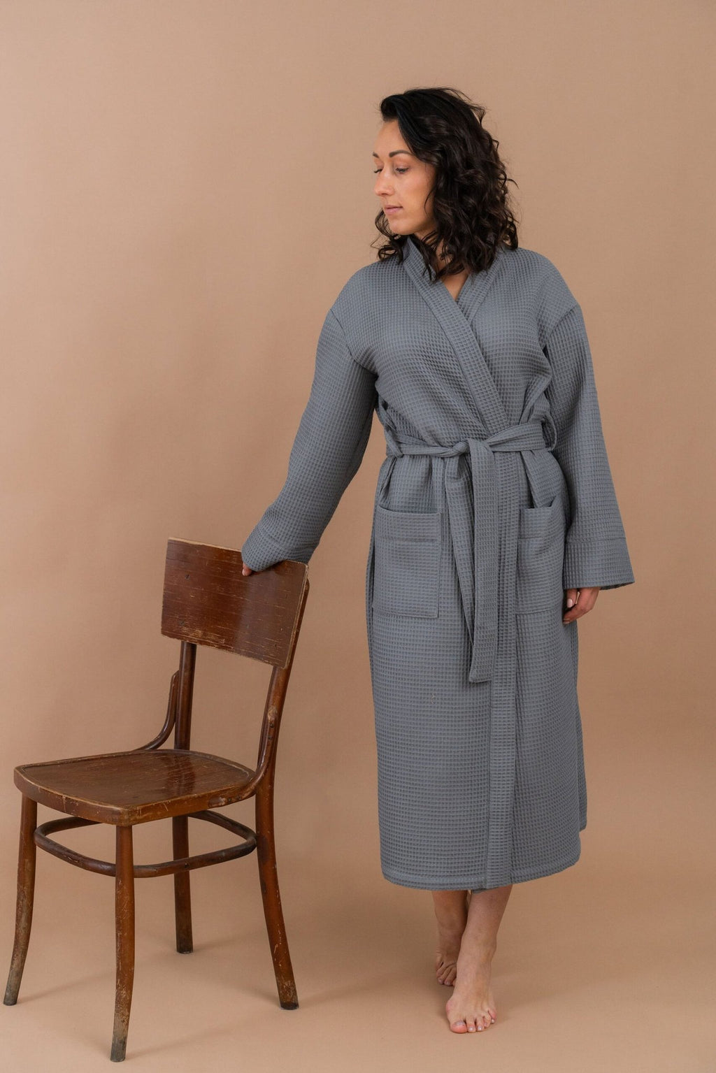 2. Woman standing barefoot in grey waffle robe with belt, next to a wooden chair, on a beige background
