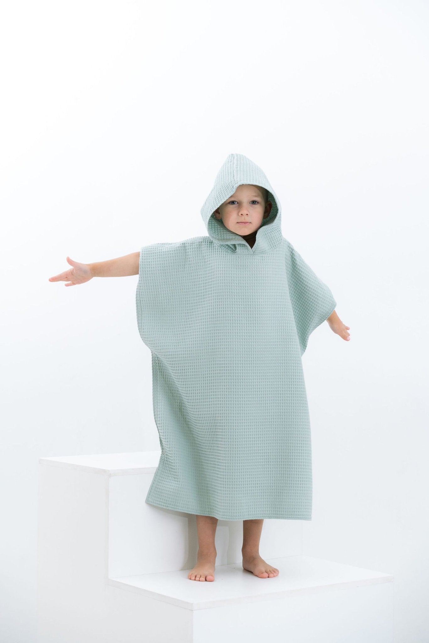 6. Child in sage green waffle fabric poncho with hood standing with arms outstretched