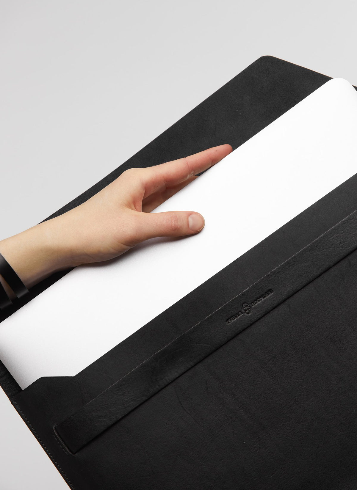 1. Hand placing a laptop into a black Stella Soomlais leather sleeve with flap, showcasing minimalist design and brand logo