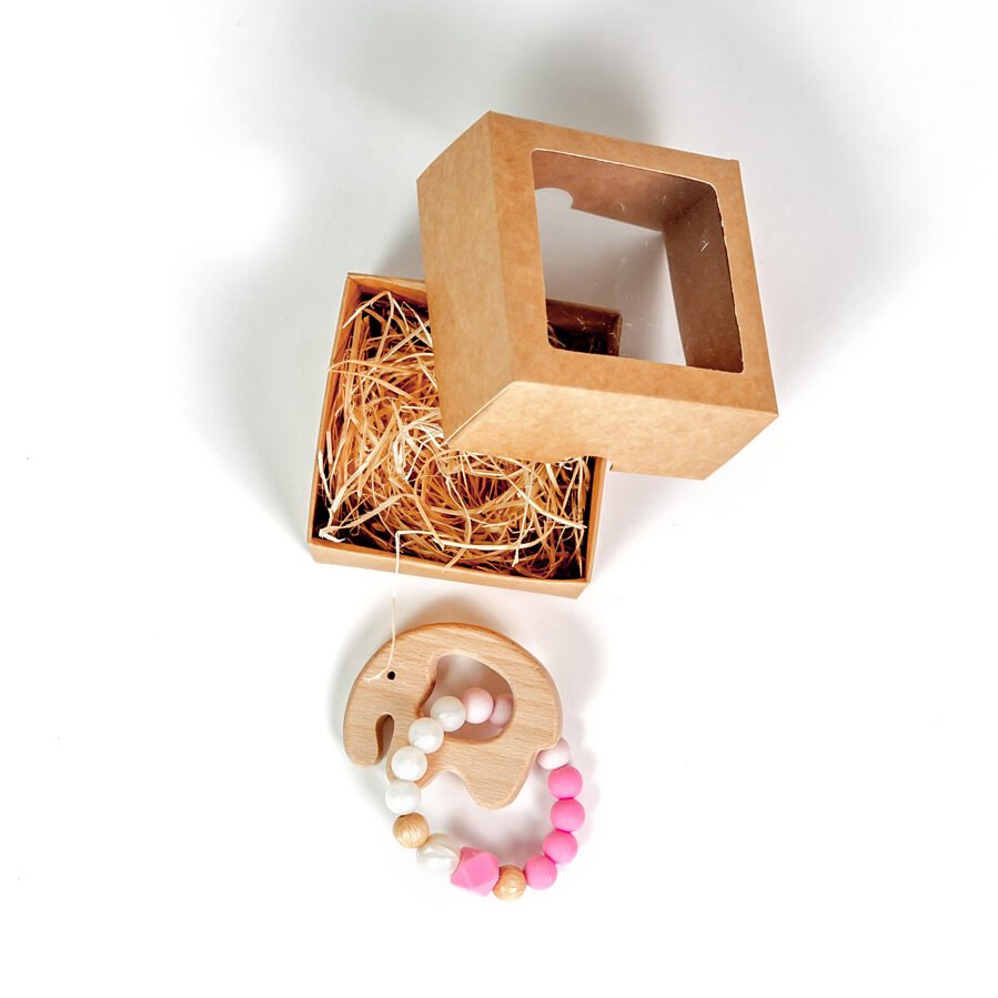 2. Light pink silicone baby teether-bracelet with wooden elephant detail, shown with open brown gift box on white background