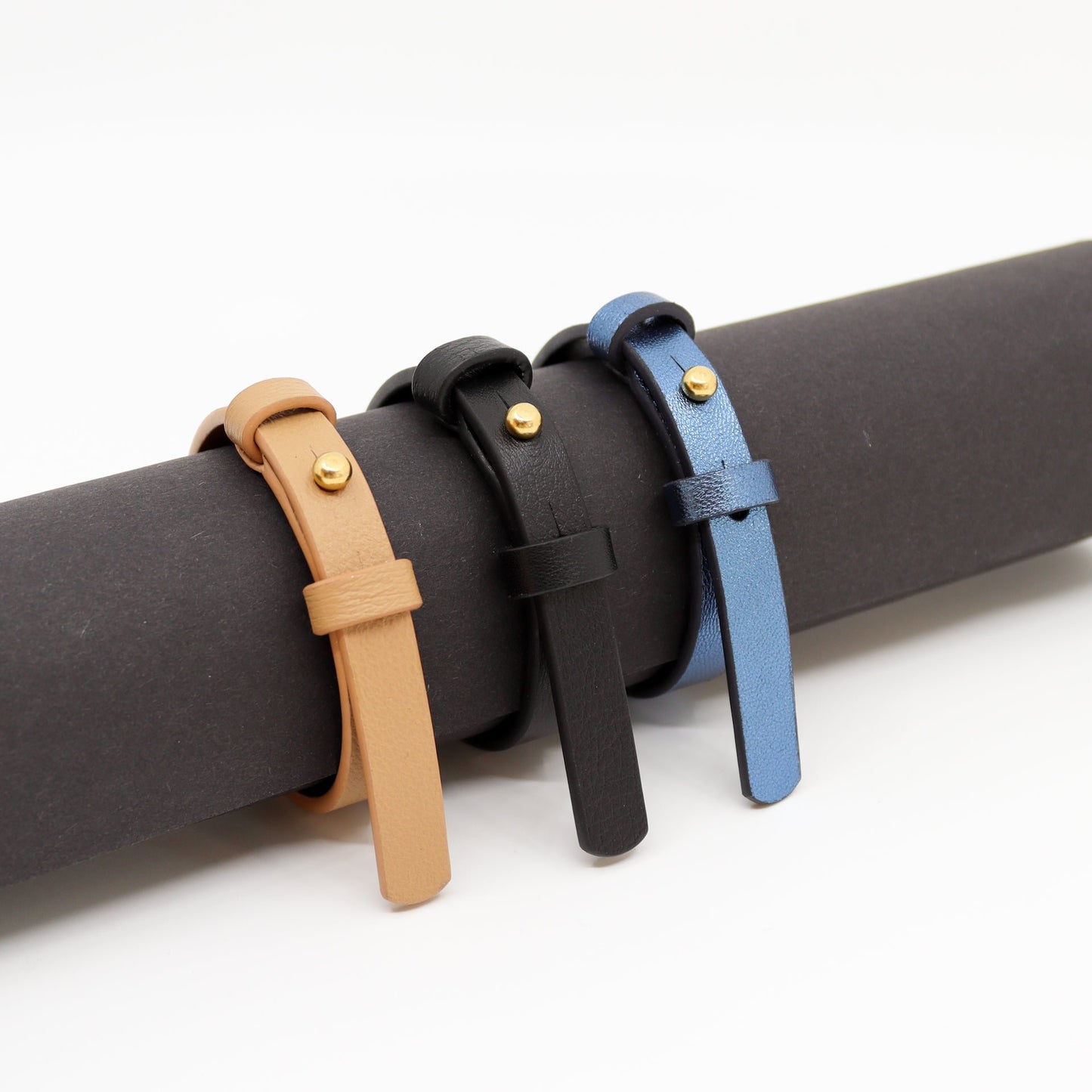2. Trio of leather bracelets in black, tan, and blue with brass fasteners displayed on a roll