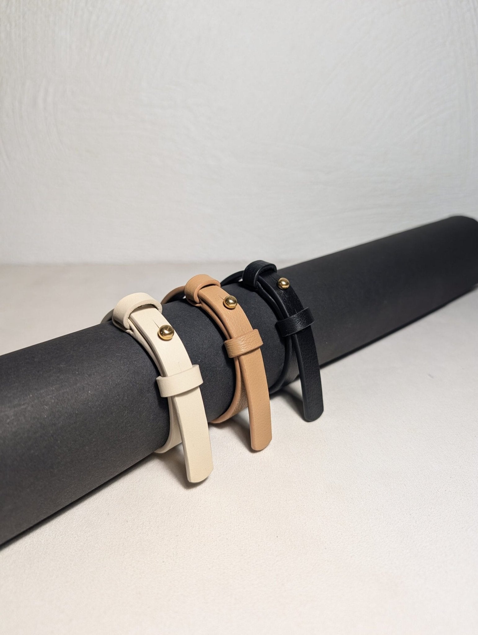4. Close-up of Tairi Roosve leather bracelets in ivory, tan, and black with brass fasteners, displayed on a roll