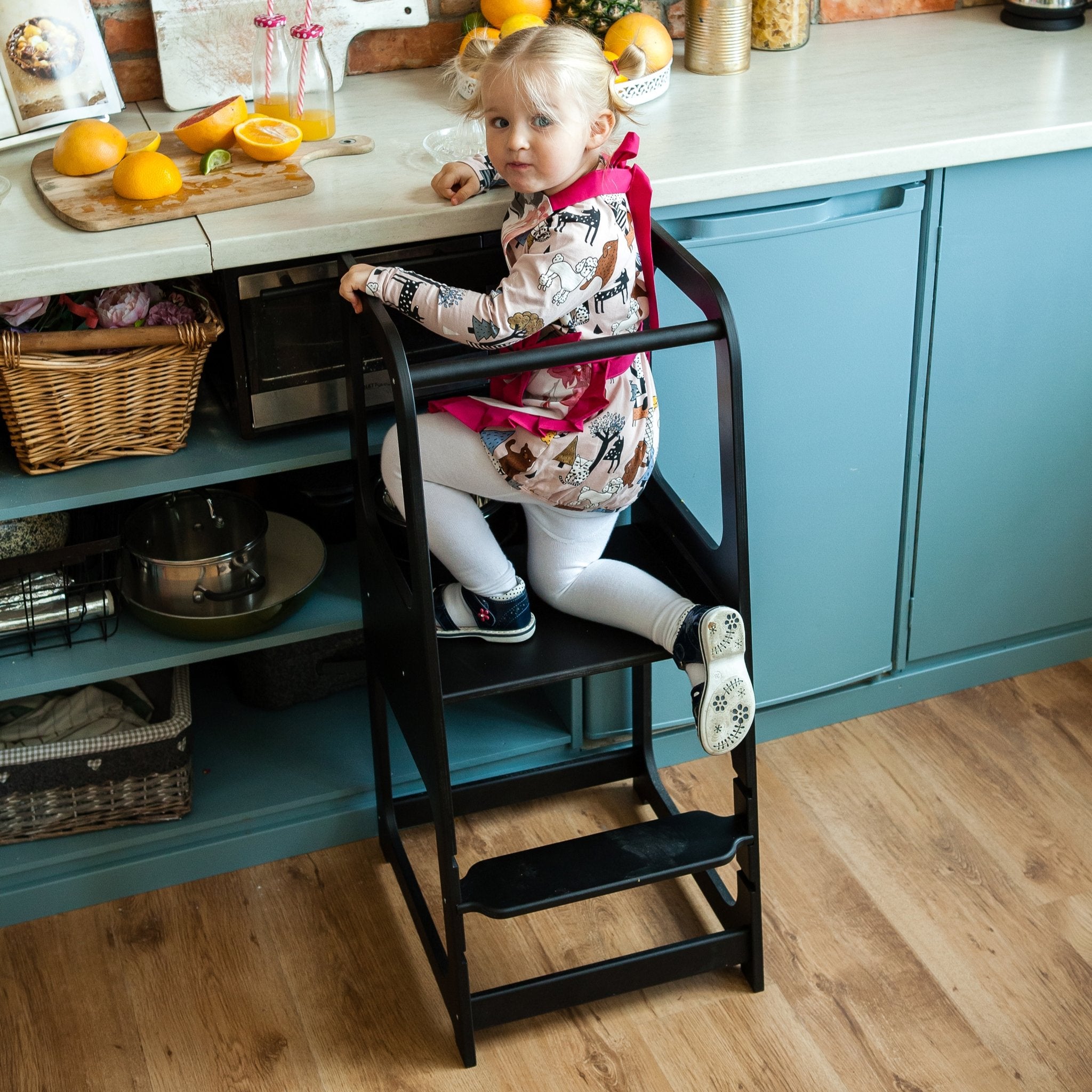 5. Child in floral dress on black Montessori helper tower step stool in kitchen