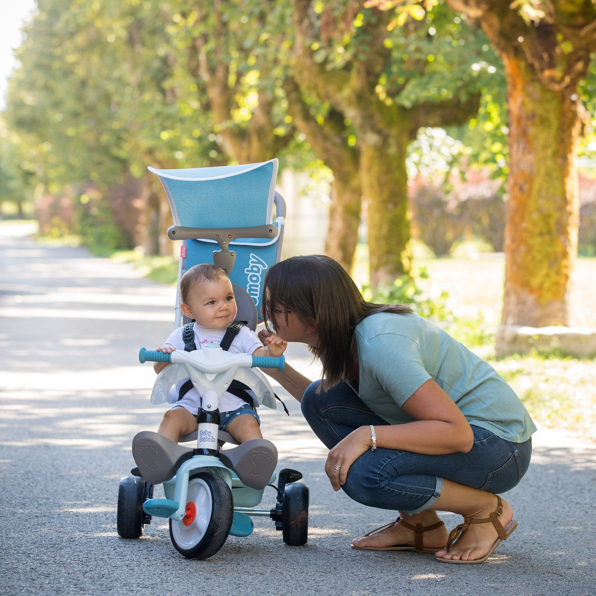 1. Woman crouching next to a child on a blue Smoby Baby Balade Plus tricycle in a sunny park