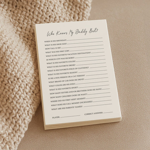 1. Stack of Beebila 'Who Knows Daddy Best' baby shower game cards on a beige knitted blanket, featuring questions about the dad-to-be on cream Munken Pure paper.