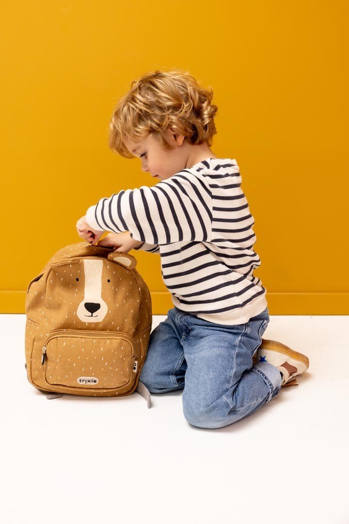 1. Child kneeling and opening Trixie Baby Mr. Bear backpack against a yellow background