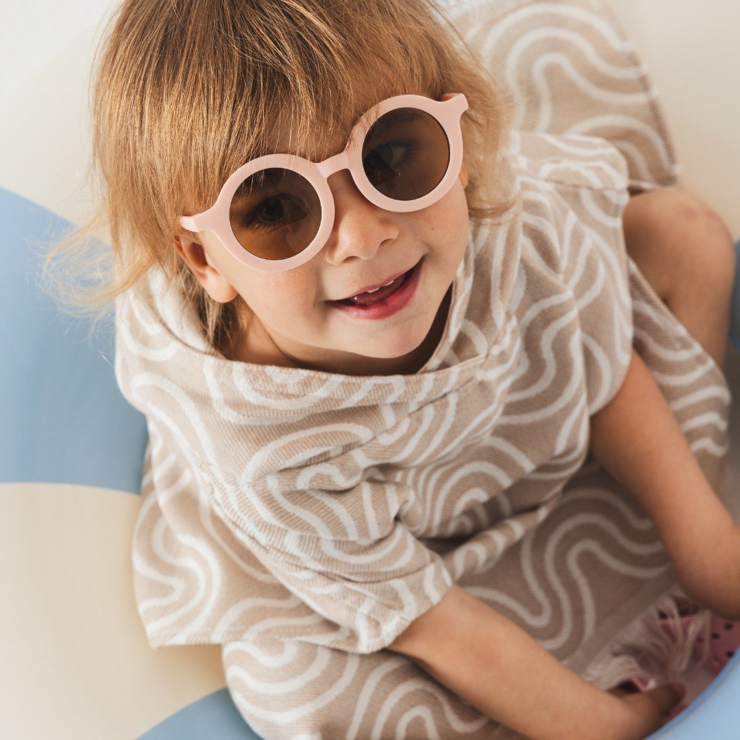 1. Child wearing sand-colored bamboo bath poncho with wave pattern and pink sunglasses sitting on inflatable pool toy