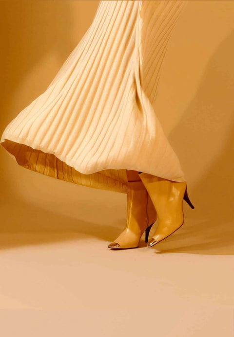 1. Woman wearing beige calf leather ankle boots with 8.7 cm heel and pointed toe, paired with a pleated skirt in a warm-toned setting