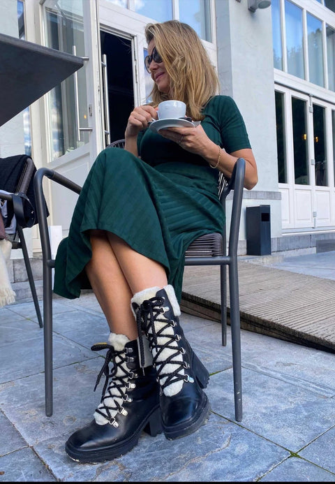 1. Woman sitting outdoors in green dress wearing black combat boots with shearling detail, holding a coffee cup