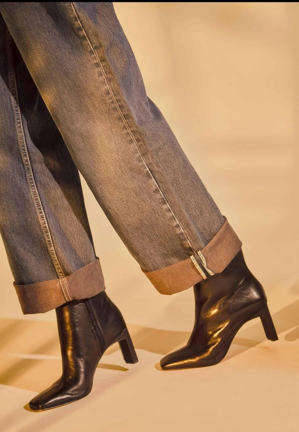 1. Person wearing black goat leather Senna boots with 7.6 cm block heel and rolled-up jeans in warm lighting