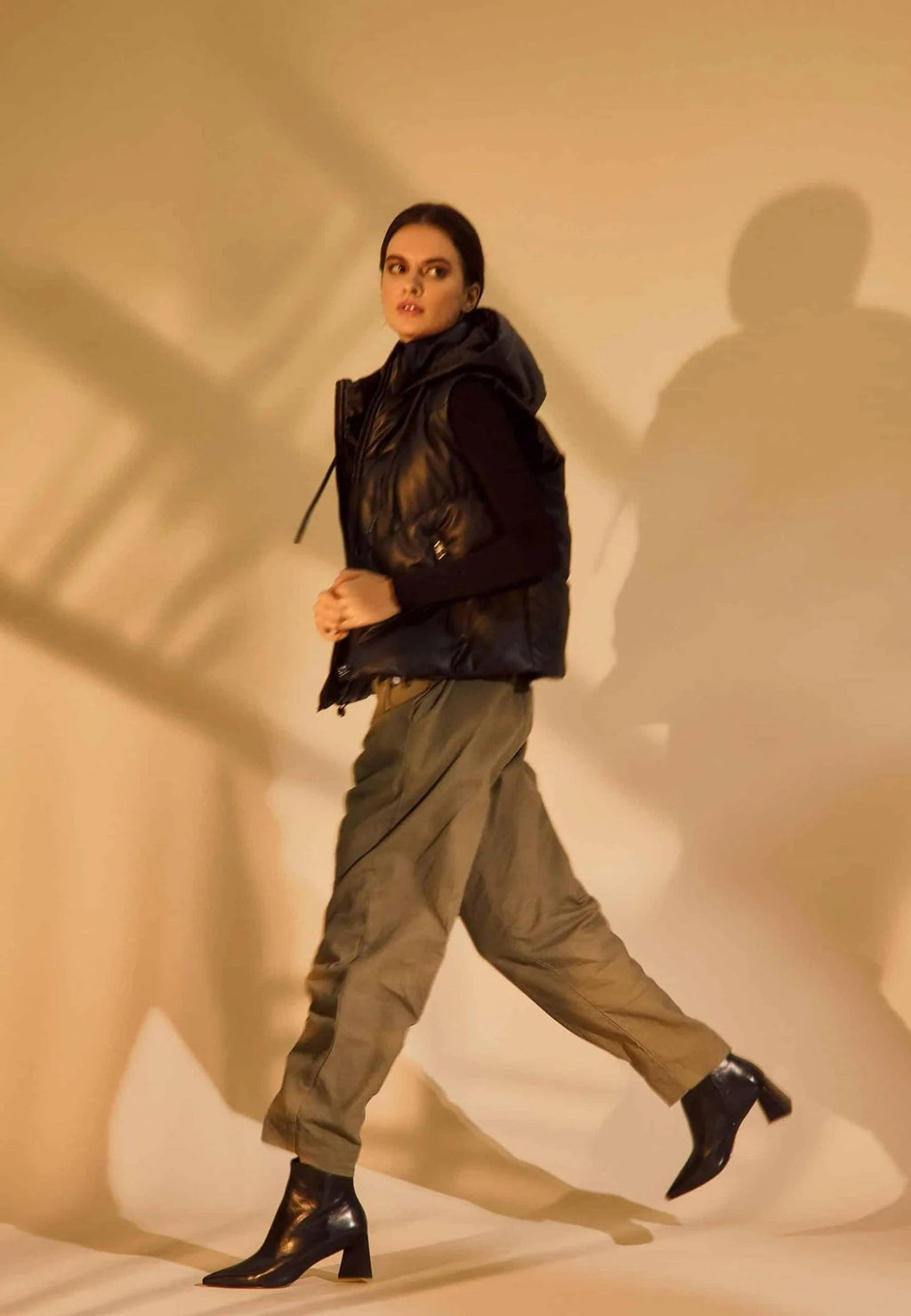 1. Woman in black vest and khaki pants wearing black leather ankle boots with pointed toe and block heel in motion on beige background