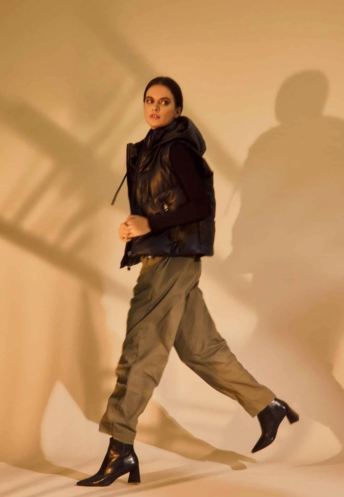1. Woman in black vest and khaki pants wearing black leather ankle boots with pointed toe and block heel in motion on beige background