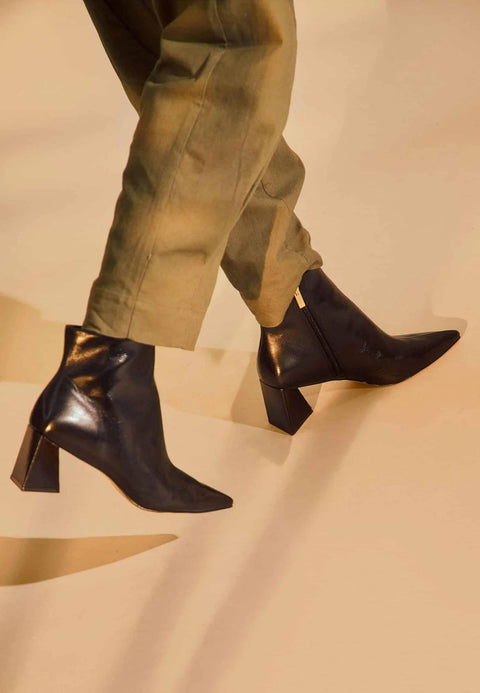 1. Woman wearing black leather ankle boots with pointed toe and block heel walking on beige background