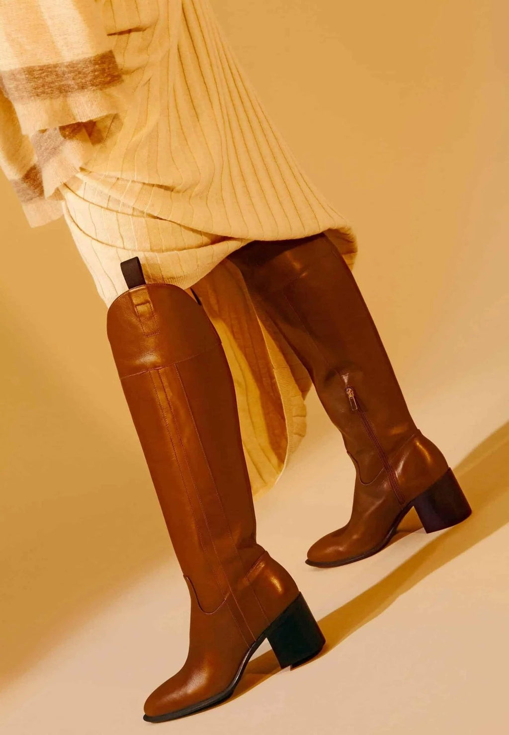 1. Woman wearing brown knee-high leather boots with block heel, styled with a beige pleated dress in a warm-toned setting
