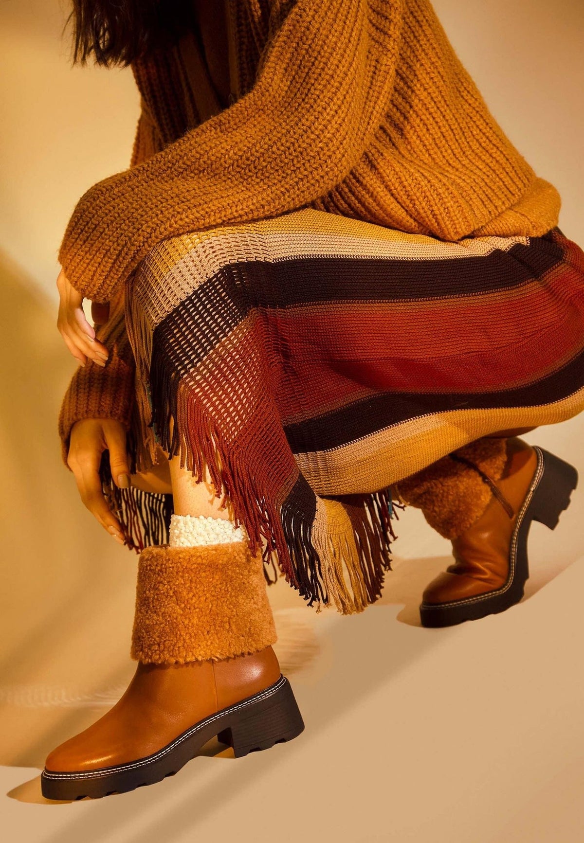 1. Woman wearing brown leather ankle boots with shearling detail, paired with a striped skirt and knit sweater in warm-toned setting